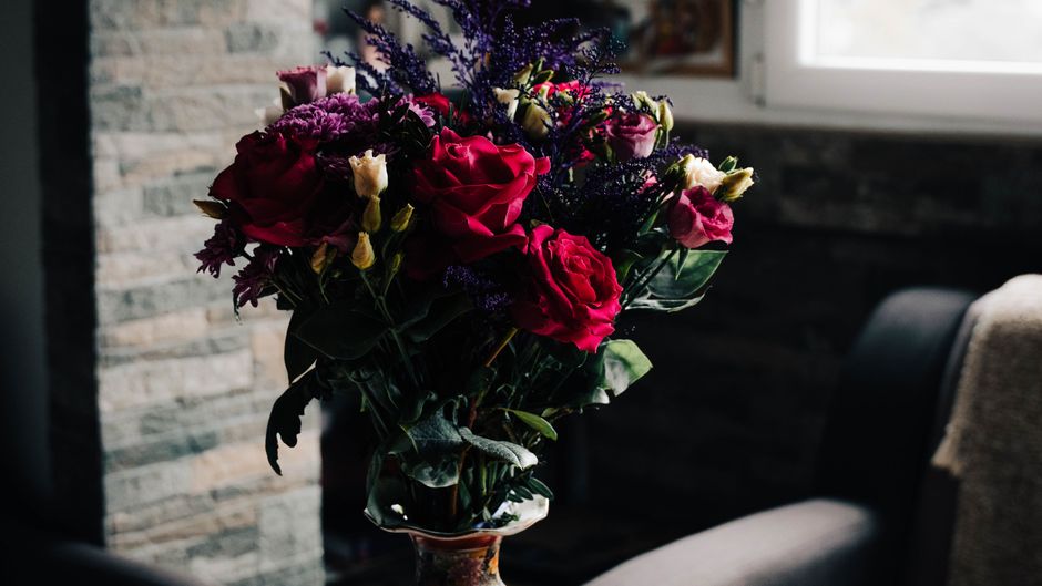 A tall floral arrangement featuring vibrant pink and deep red roses, purple statice, and white lisianthus blooms, arranged in a clear glass vase with green foliage. The bouquet is placed on a table in