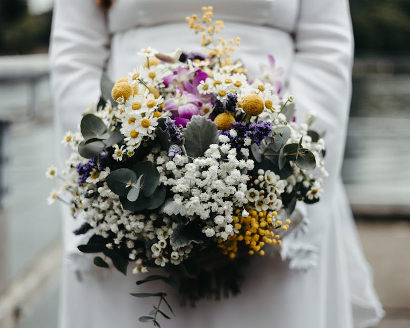 A person dressed in a white outfit is holding a large, lush bouquet featuring a variety of fresh flowers, including white daisies with yellow centers, purple orchids, small white baby's breath, yellow