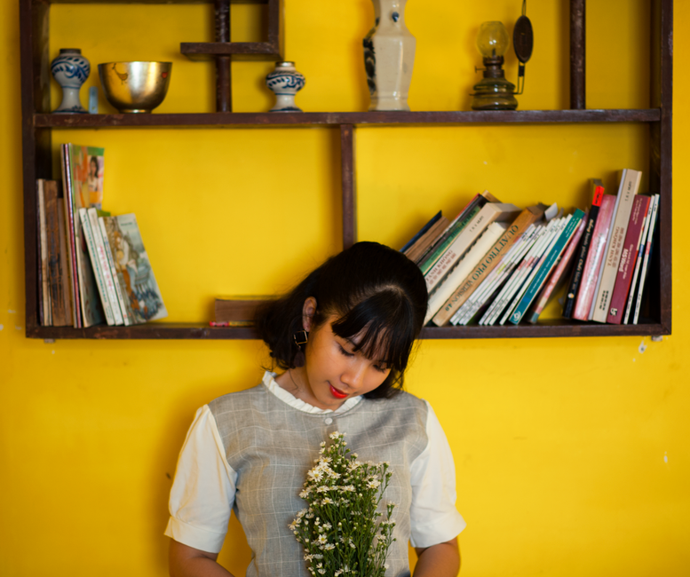 A young woman with dark hair and bangs, wearing a light grey checkered dress with white sleeves, is looking down at a bouquet of small white flowers she is holding gently in front of her. She is stand