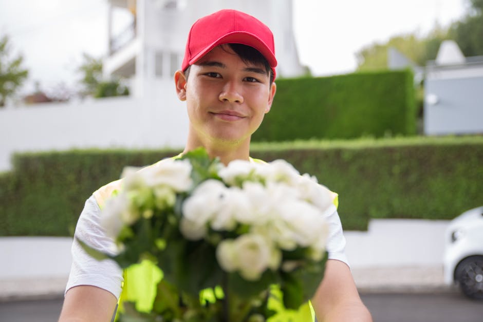 A delivery person wearing a red baseball cap and a high-visibility vest holds out a bouquet of fresh white flowers, possibly roses or ranunculus, wrapped in clear floral wrap. The arrangement features